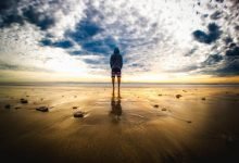 A lone figure stands on a tranquil beach during a vivid sunset, reflecting on the wet sand.