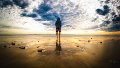 A lone figure stands on a tranquil beach during a vivid sunset, reflecting on the wet sand.